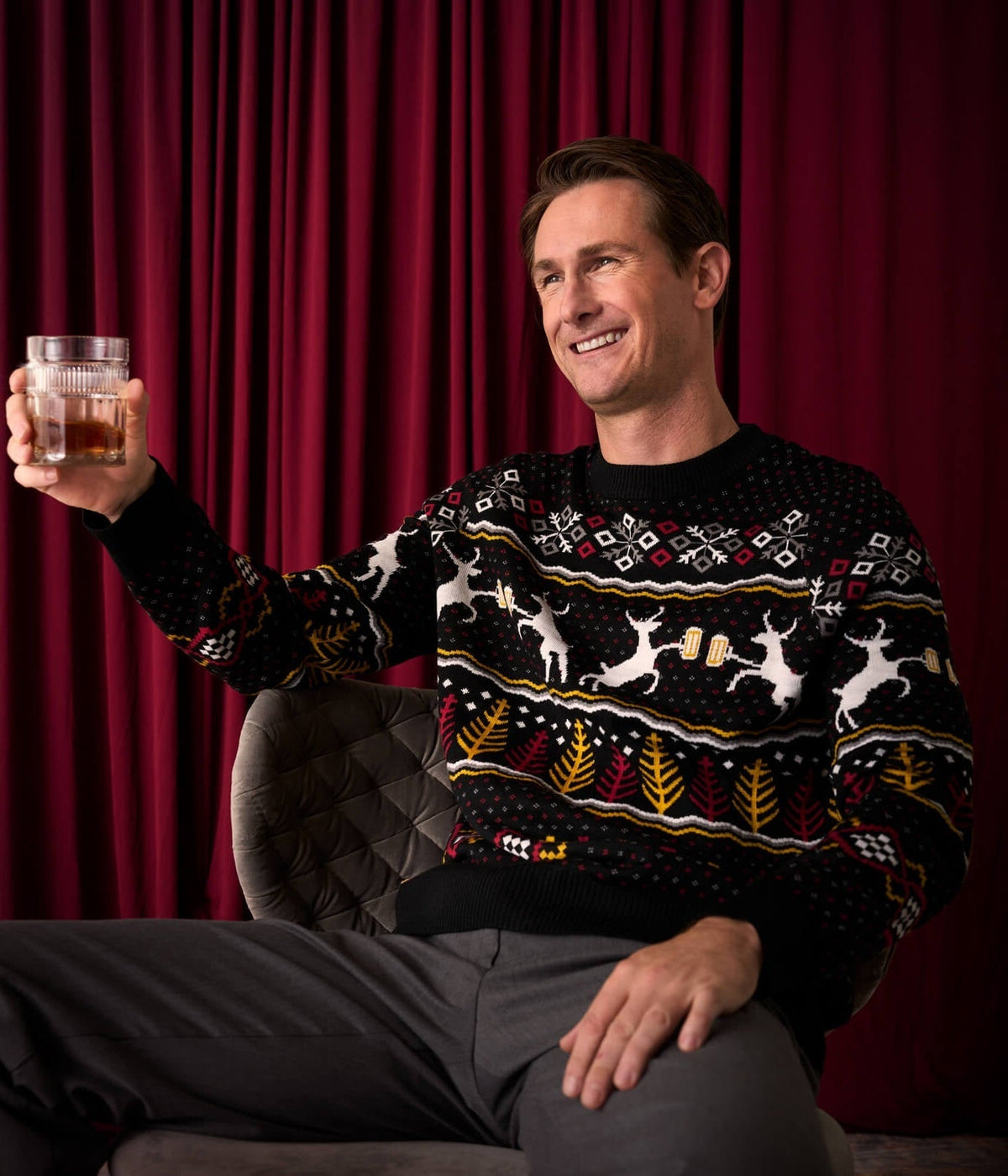 Man wearing a festive sweater holding a glass of whiskey against a red curtain background