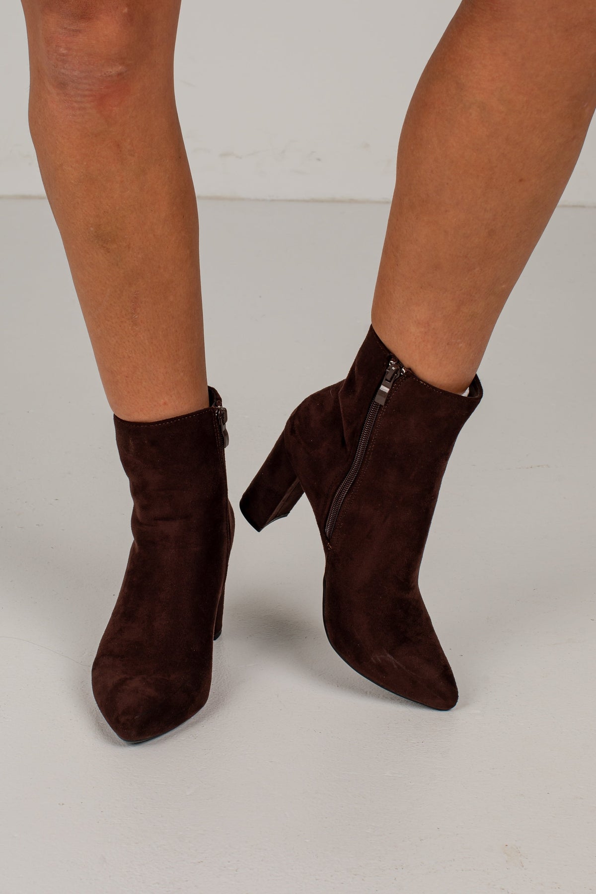 A pair of dark brown suede ankle boots with pointed toes and block heels, shown on a model's feet against a light gray studio background.