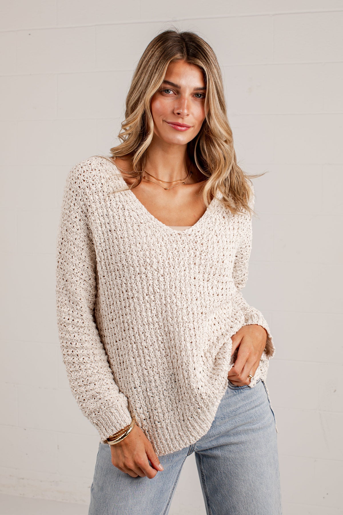A woman wearing a chunky knit cream-colored sweater and light blue jeans, standing against a white paneled wall.